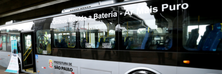 Fotografia da lateral de um ônibus nas cores prata e preto, dando para ver os bancos azuis com amarelo no interior através dos vidros das janelas.